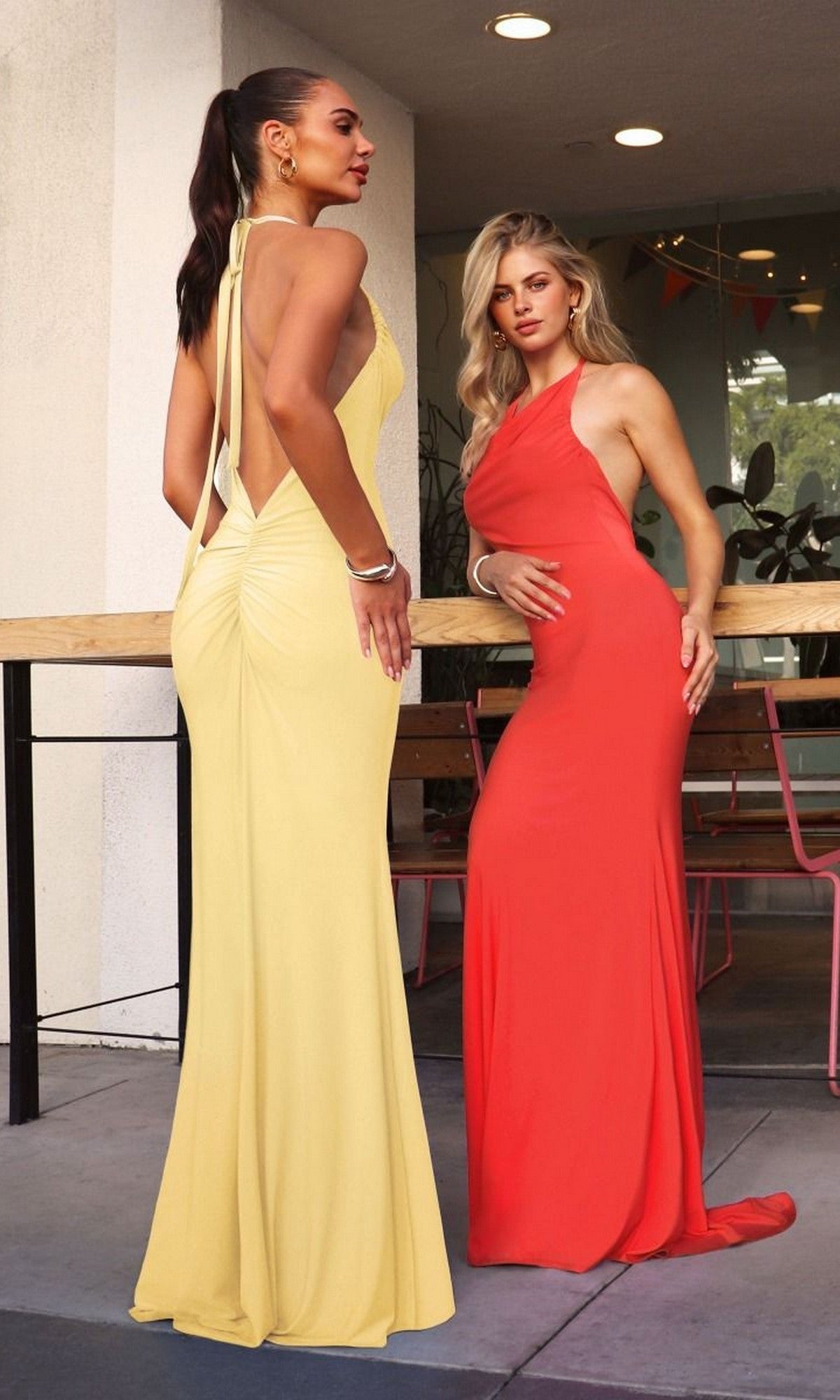 Two women stand outside near a wooden table, one in the Astee T506 long prom dress in yellow with a low back and the other in an orange gown with a convertible halter neckline.